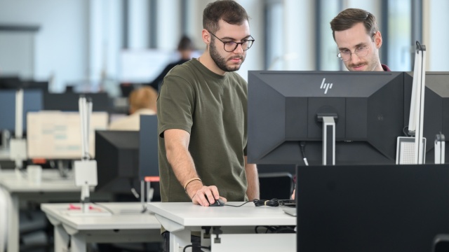 Két férfi áll egy monitor előtt, ami egy irodai asztalon áll egy irodai környezetben
