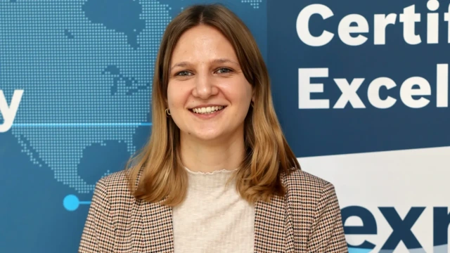 A woman with shoulder-length hair is smiling while standing in front of a blue backdrop featuring text and a graphic world map. She is wearing a checkered blazer over a light shirt.