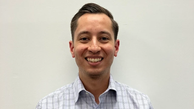 A man with short, dark hair is smiling at the camera. He is wearing a plaid shirt against a neutral white background.