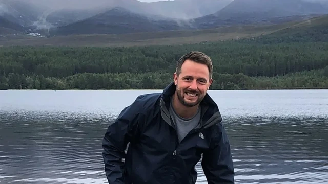 A man in a dark jacket stands by a lake with green hills and misty mountains in the background.