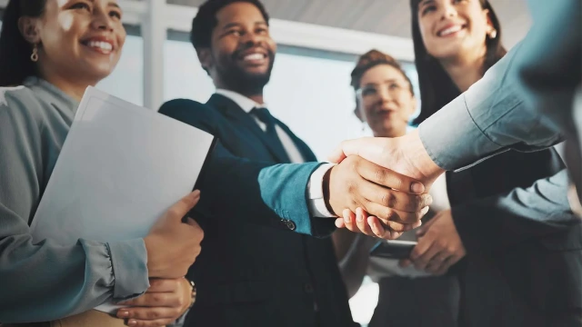 Professionals smiling and shaking hands in a modern office, indicating a successful business agreement. Three people in the background, one holding documents.