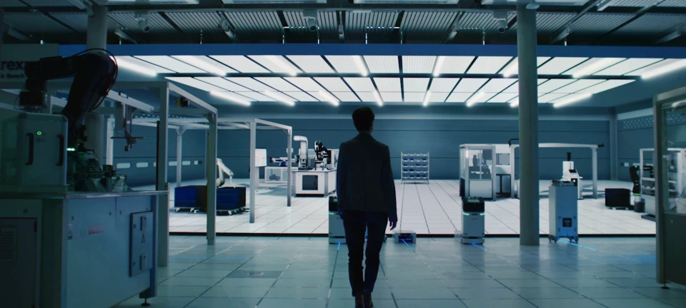 person walking towards a modern, industrial-looking workspace filled with advanced machinery and robotics. The room is well-lit with overhead lighting panels, and the environment has a clean, high-tech appearance.