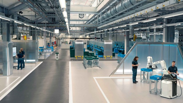 Menschen, AGV und Roboter arbeiten gemeinsam in einer modernen, industriellen Werkhalle.