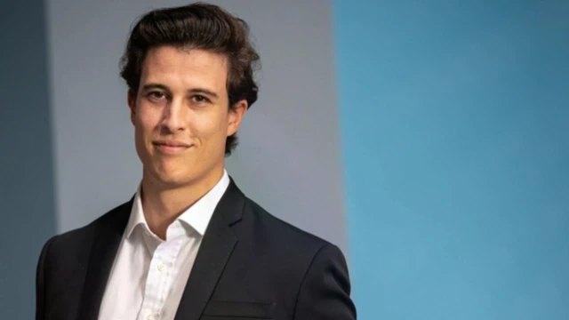 A young man with brown hair and brown eyes wearing a collared shirt and a blazer is smiling.