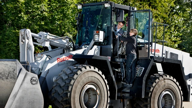 Two people, one man and one woman are operating a grey excavator with a Rexroth logo on it.