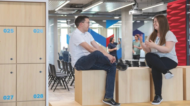 Two people with t-shirts, one man and one woman are sitting on blocks and talking to each other. They are in a brightly lit office setting.