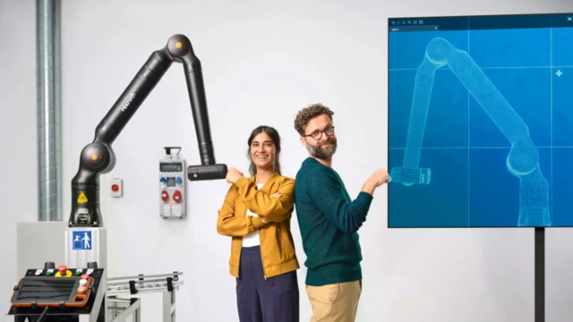 A woman and a man stand back to back. The woman is fist-bumping a robot while the man is fist-bumping a digital twin of that robot.