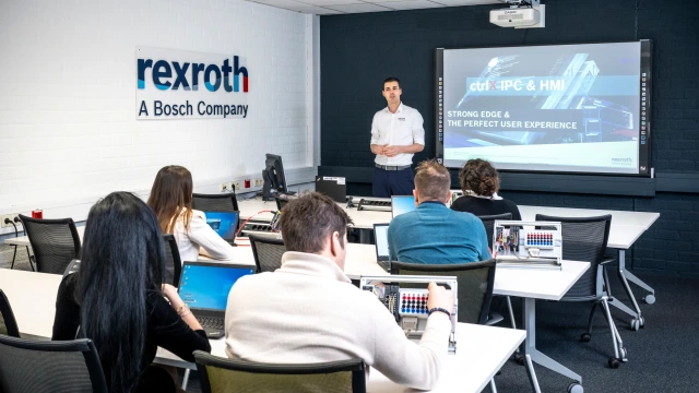 A group of diverse people are sitting in a class-room like setting and listening to a man who is upfront talking. The Rexroth logo can be seen on the wall.