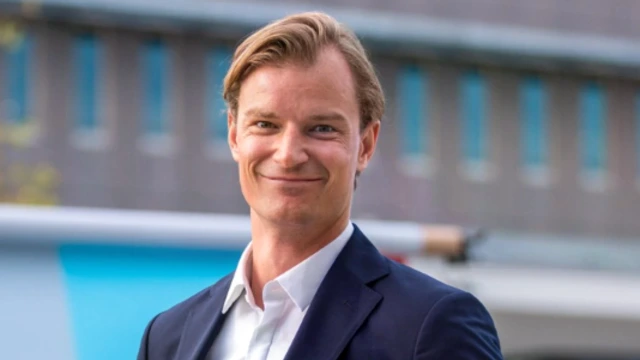 A young white man with blond hair and brown eyes is smiling into the camera. He is outside and an office building is behind him.