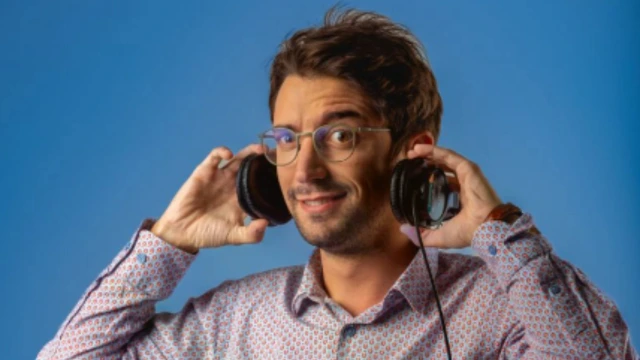 A young man with glasses is taking off his black headphones. The wall behind him is blue and he is wearing a patterned long-sleeved collared shirt.