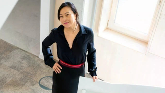 A woman of asian decent wearing a black dress is standing next to a staircase in a modern office setting.