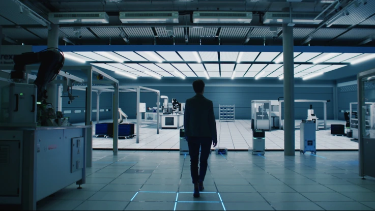 person walking towards a modern, industrial-looking workspace filled with advanced machinery and robotics. The room is well-lit with overhead lighting panels, and the environment has a clean, high-tech appearance.