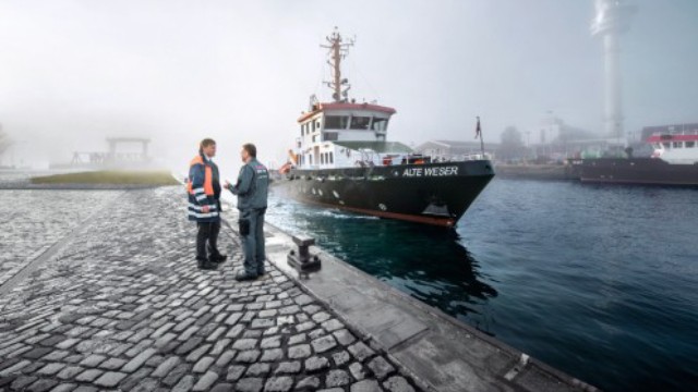Zwei Hafenmitarbeiter stehen am Kai während ein Schiff einläuft.