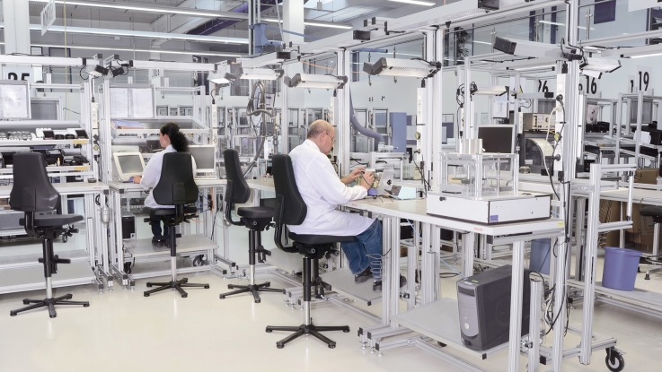 Several manual workstations and feeding systems, arranged in an L-shape. 4 chairs, 2 of which are occupied by workers. In the background, roll feeders and returns can be seen. The workstations are equipped with gripping containers, work equipment, information boards, and material supply using shelving systems and gripping containers.