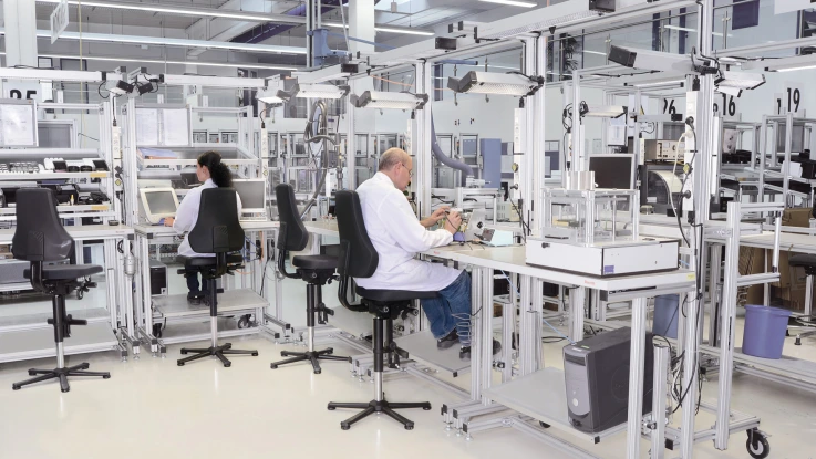 Several manual workstations and feeding systems, arranged in an L-shape. 4 chairs, 2 of which are occupied by workers. In the background, roll feeders and returns can be seen. The workstations are equipped with gripping containers, work equipment, information boards, and material supply using shelving systems and gripping containers.