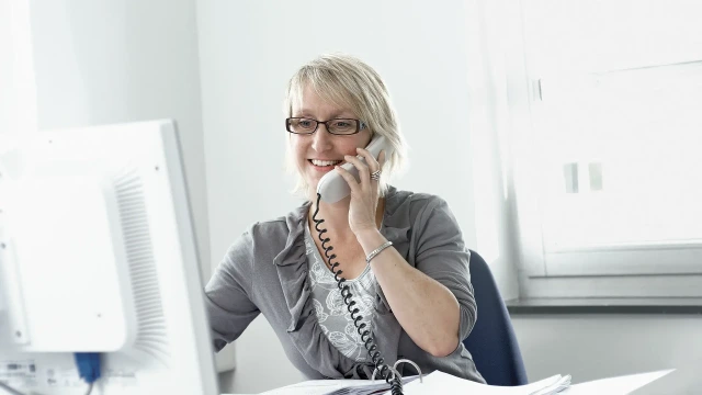 The picture shows a woman at a desk with a monitor talking on the phone.