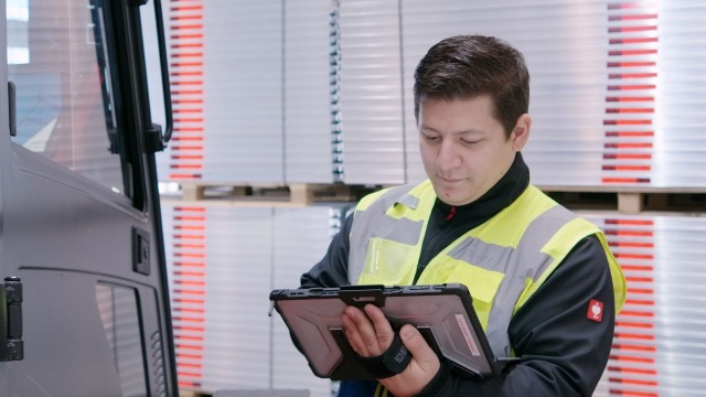 De afbeelding toont een magazijnmanager die een tablet bedient. Links van hem staan delen van een vorkheftruck. Achter hem staan steigerelementen.