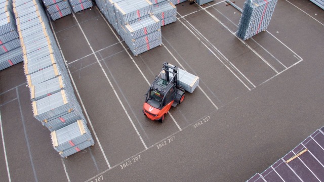 L’image montre un chariot élévateur chargé de matériel (éléments d’échafaudage) dans un entrepôt en extérieur.