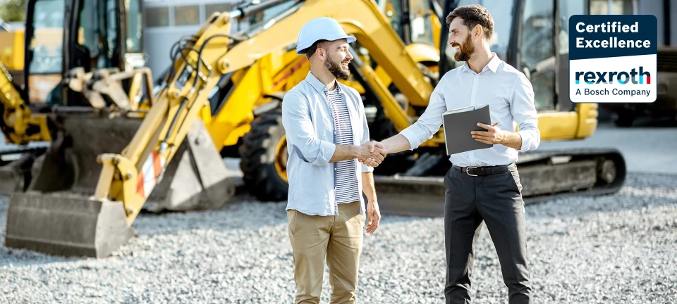 Partener și client final care își strâng mâna în fața unui excavator