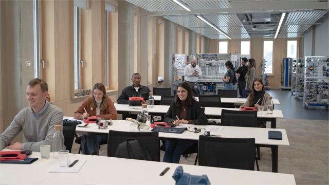 Elever sitter ved bord. XITE Hydraulix 300-opplæringssystemet fra Bosch Rexroth i bakgrunnen