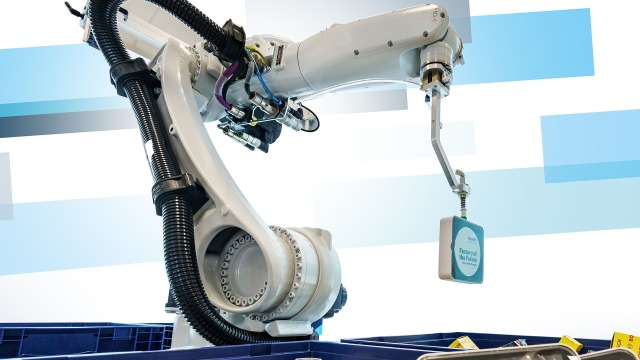 Robot arm surrounded by boxes filled with objects. The arm grabs an object that says, "Factory of the future".