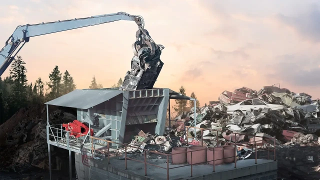 The picture shows a crane lifting a car wreck into a pre-shredder, powered by a Hägglunds motor, at a scrap yard as the sun sets.